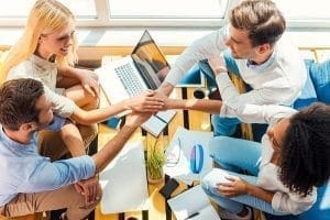 good-job-team-top-view-four-happy-young-people-holding-hands-clasped-while-sitting-wooden-desk-together - Kickstart Bahrain - Kickstart Bahrain